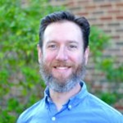 Smiling bearded man in a blue button-down shirt outdoors, with greenery and a brick wall blurred in the background.